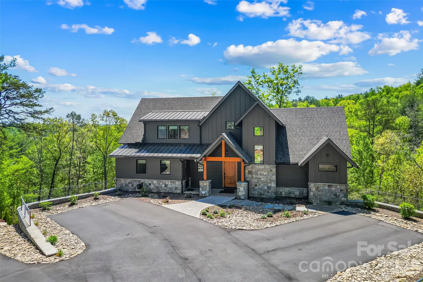 Modern two-story house with gray siding, stone accents, and a dark gray roof, surrounded by green trees under a blue sky.