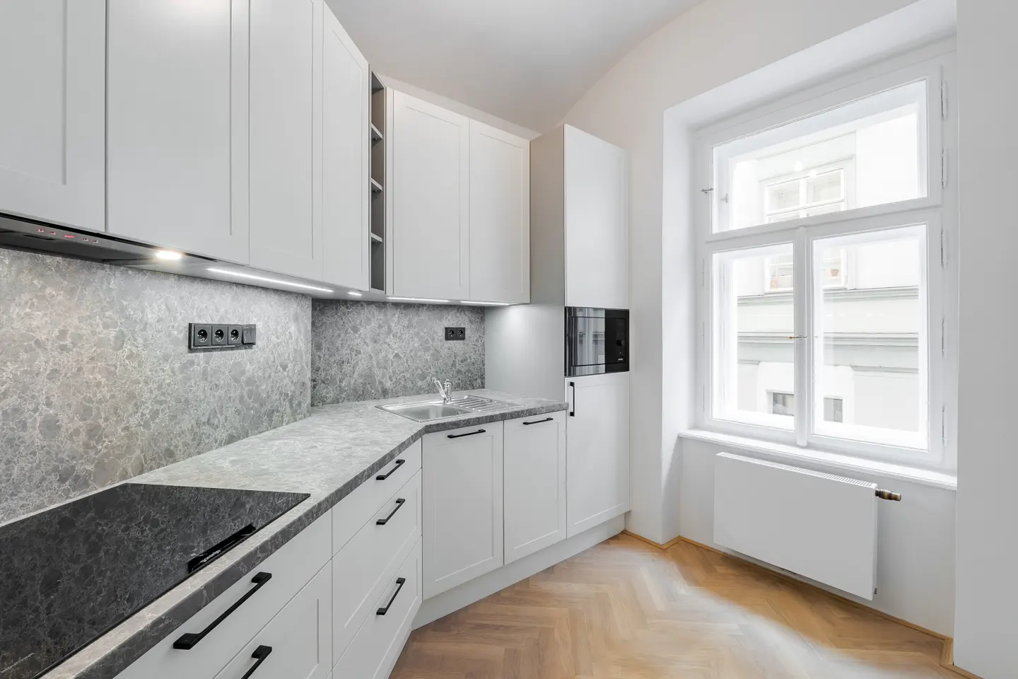 Bright kitchen with white cabinets, gray countertops and backsplash, and a herringbone wood floor. A large window provides natural light.
