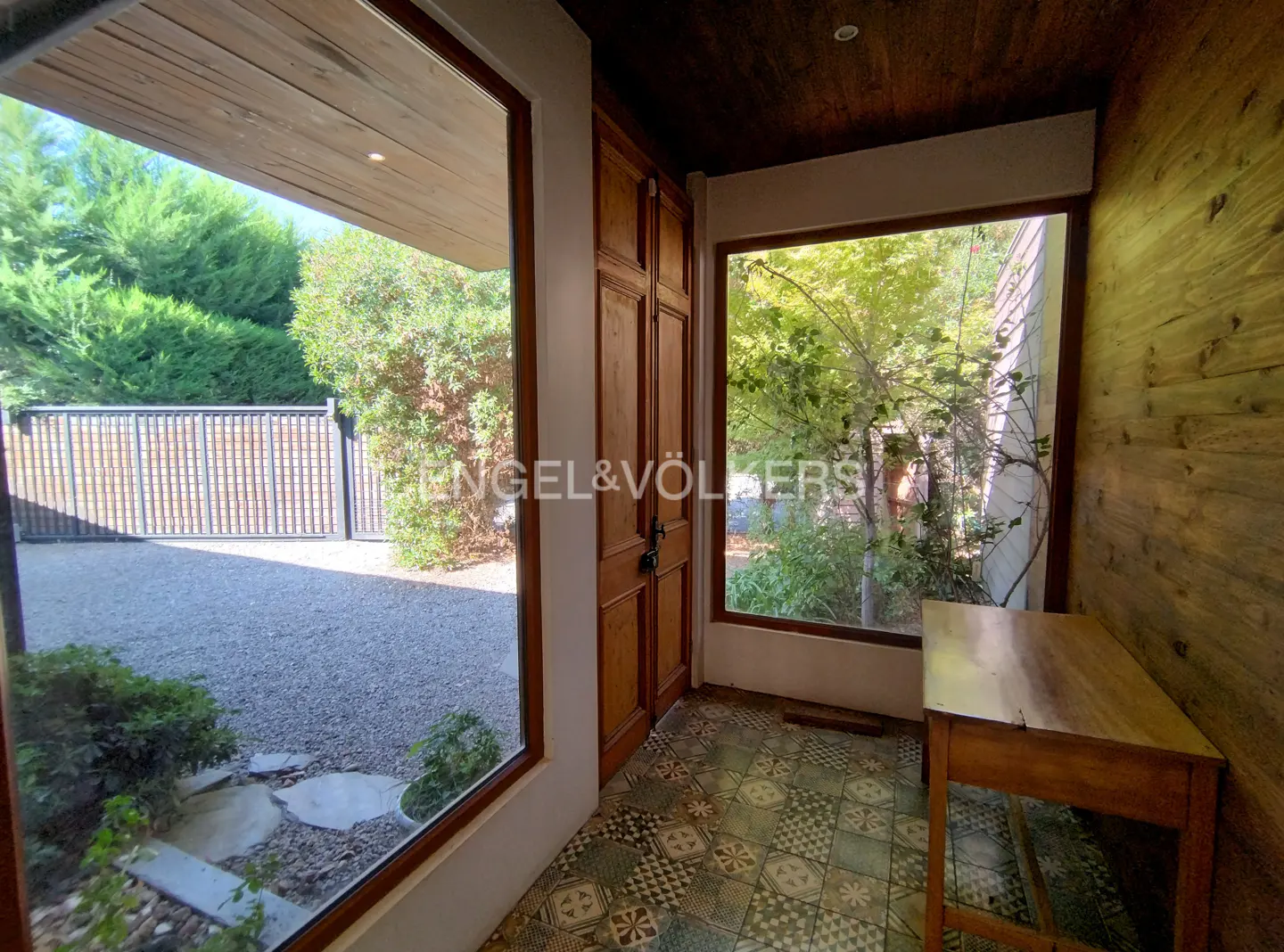 Entryway with wood door, patterned tile floor, and wood walls. Windows show greenery outside. A small wood table sits to the right.