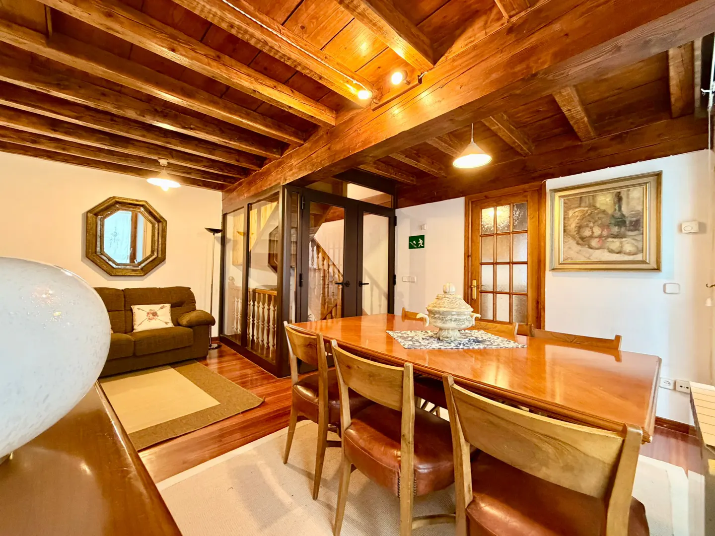 Dining room with wooden table, leather chairs, and exposed beam ceiling, adjacent to a living area with a brown sofa.