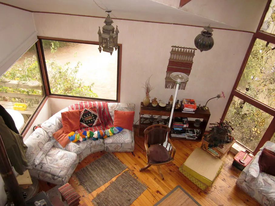 A cozy living room with a patterned sofa, colorful pillows, and wooden floors. Large windows offer a view of greenery. Decorative lamps hang from the ceiling.