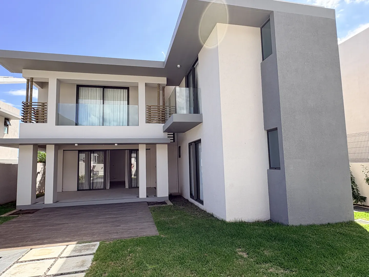 Two-story modern house with white and gray exterior, glass balcony, and open patio on a green lawn under a blue sky.