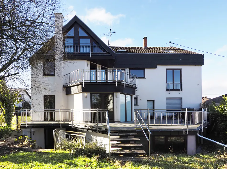 Exterior of a modern white house with black trim, multiple balconies, and a large deck with stairs leading to the yard.
