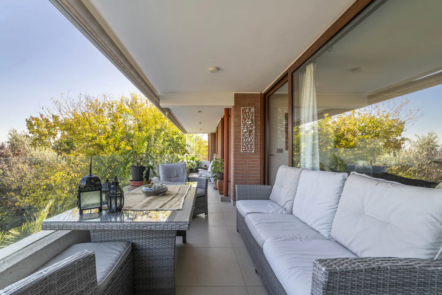 A long, covered balcony with gray wicker furniture and a view of green trees.