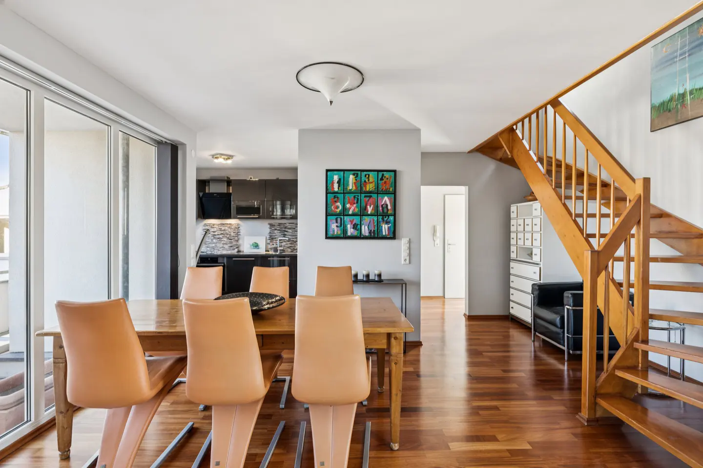 A modern dining room with a wooden table, tan chairs, and a staircase leading to the upper level. The kitchen is visible in the background.