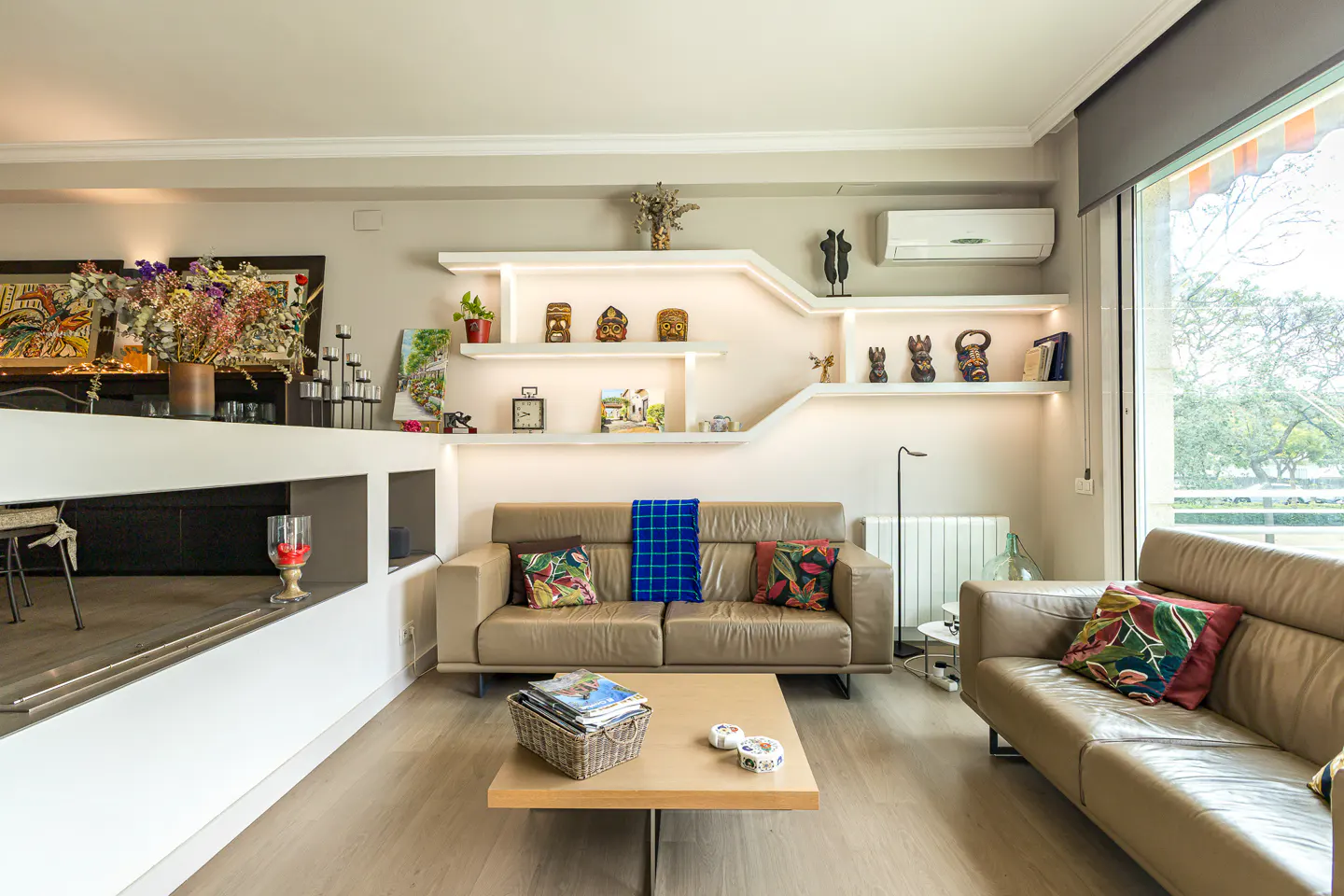 Living room with beige sofas, a light wood floor, and built-in shelves with decorative items. Large window on the right.