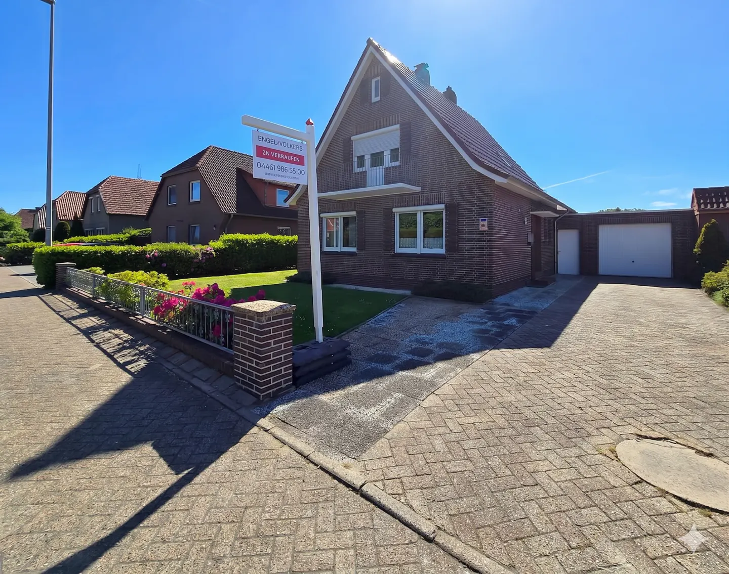 A brick house with a for-sale sign, a paved driveway, and a small front garden under a clear blue sky.