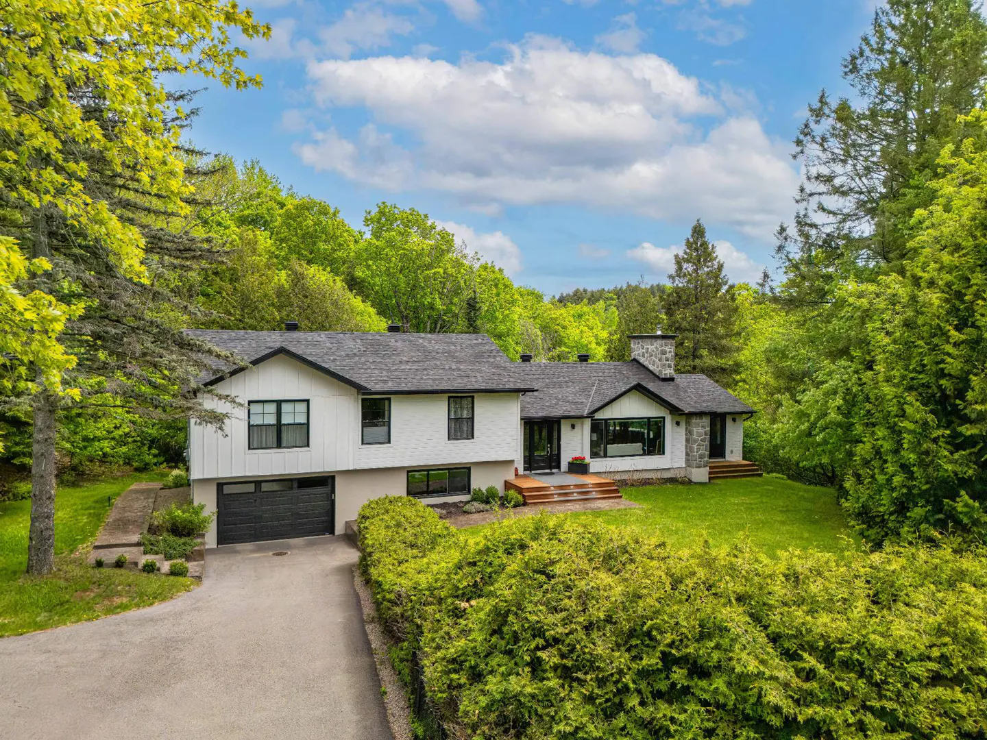 White two-story house with a dark roof and garage, surrounded by lush green trees and a lawn under a blue sky.