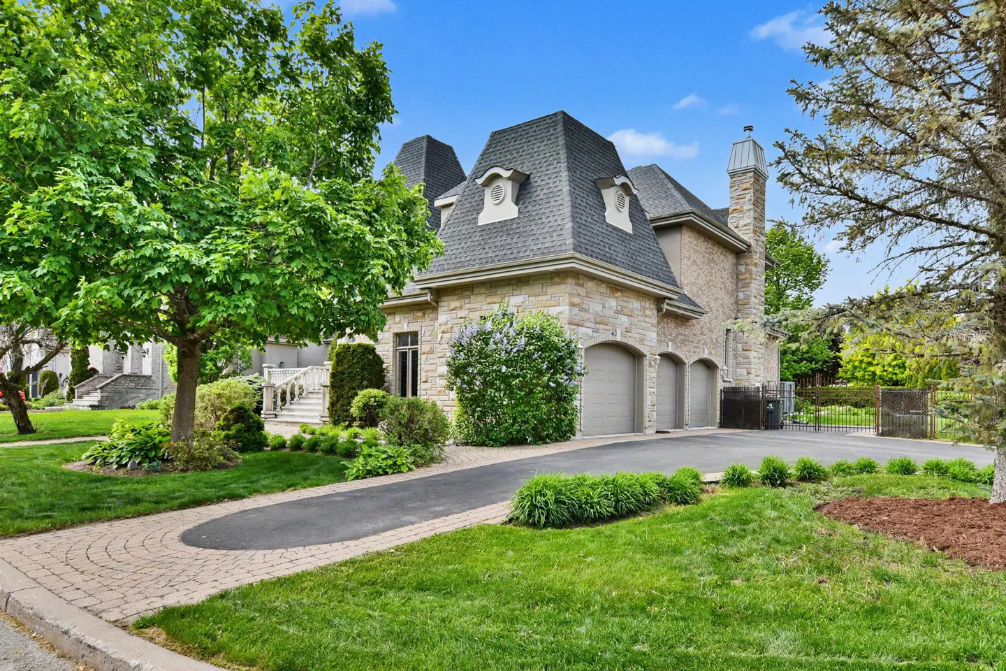 Exterior view of a stone house with a gray roof, three garage doors, and a paved driveway surrounded by green grass and trees.
