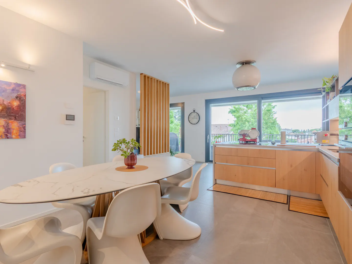 Bright, modern kitchen and dining area with white oval table, chairs, and wood cabinets. Large windows offer a view of greenery.