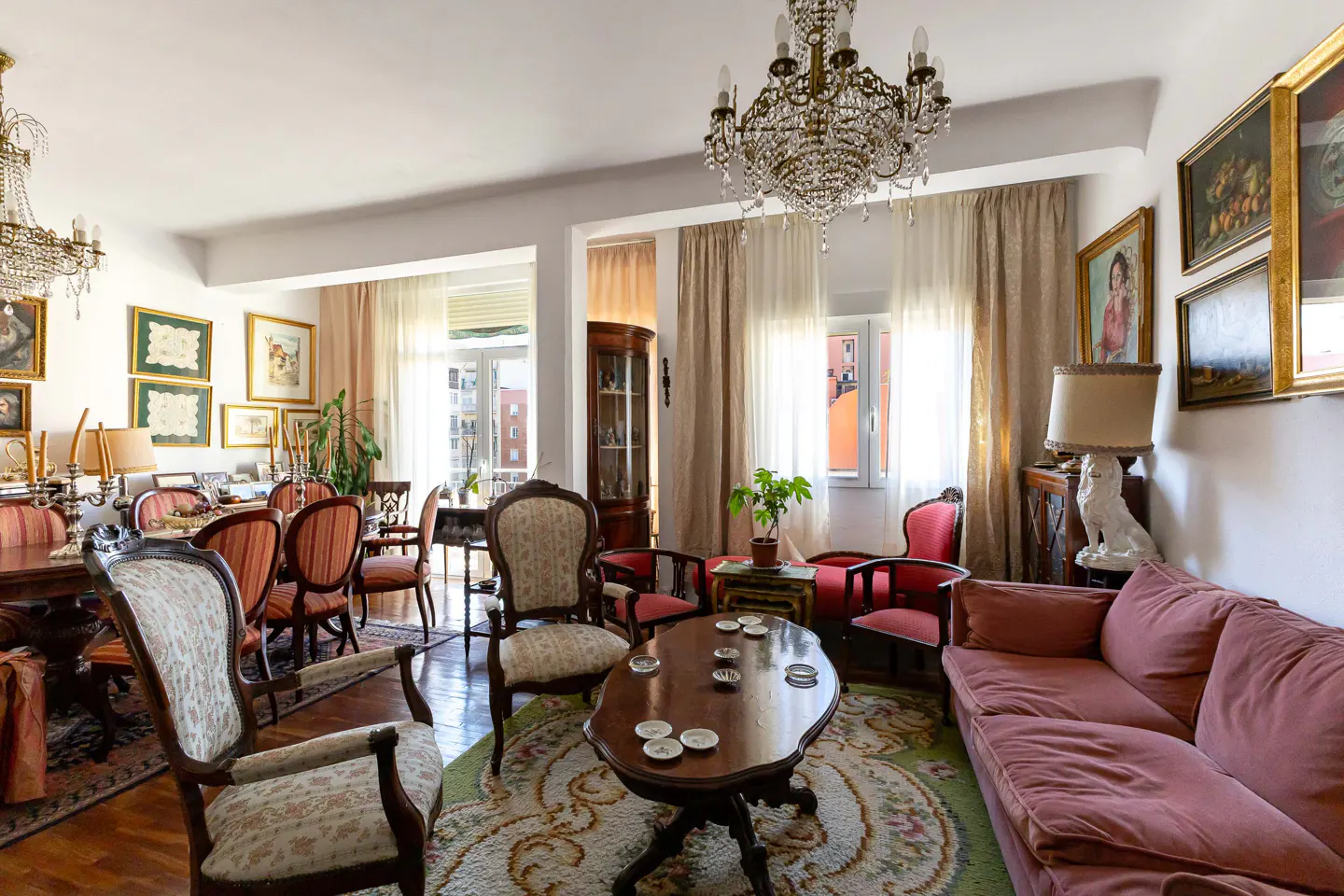 A living room with a pink sofa, wooden chairs, a table, and a crystal chandelier. Paintings adorn the walls.