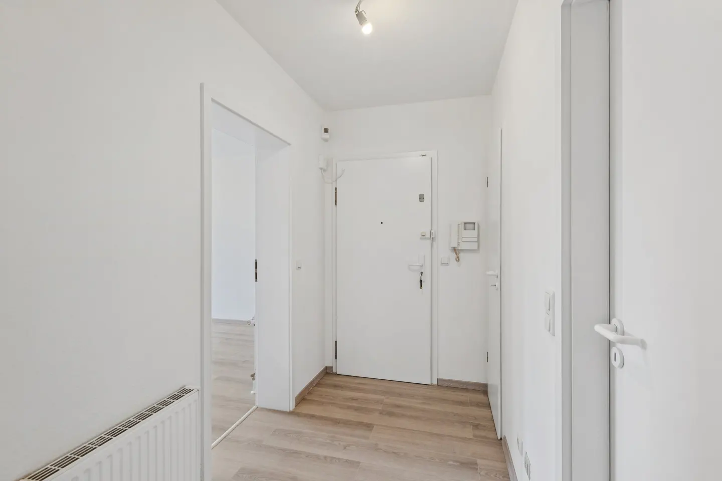 Bright, white hallway with light wood floors. A white front door is centered, with an open doorway to the left. A radiator is visible on the left wall.