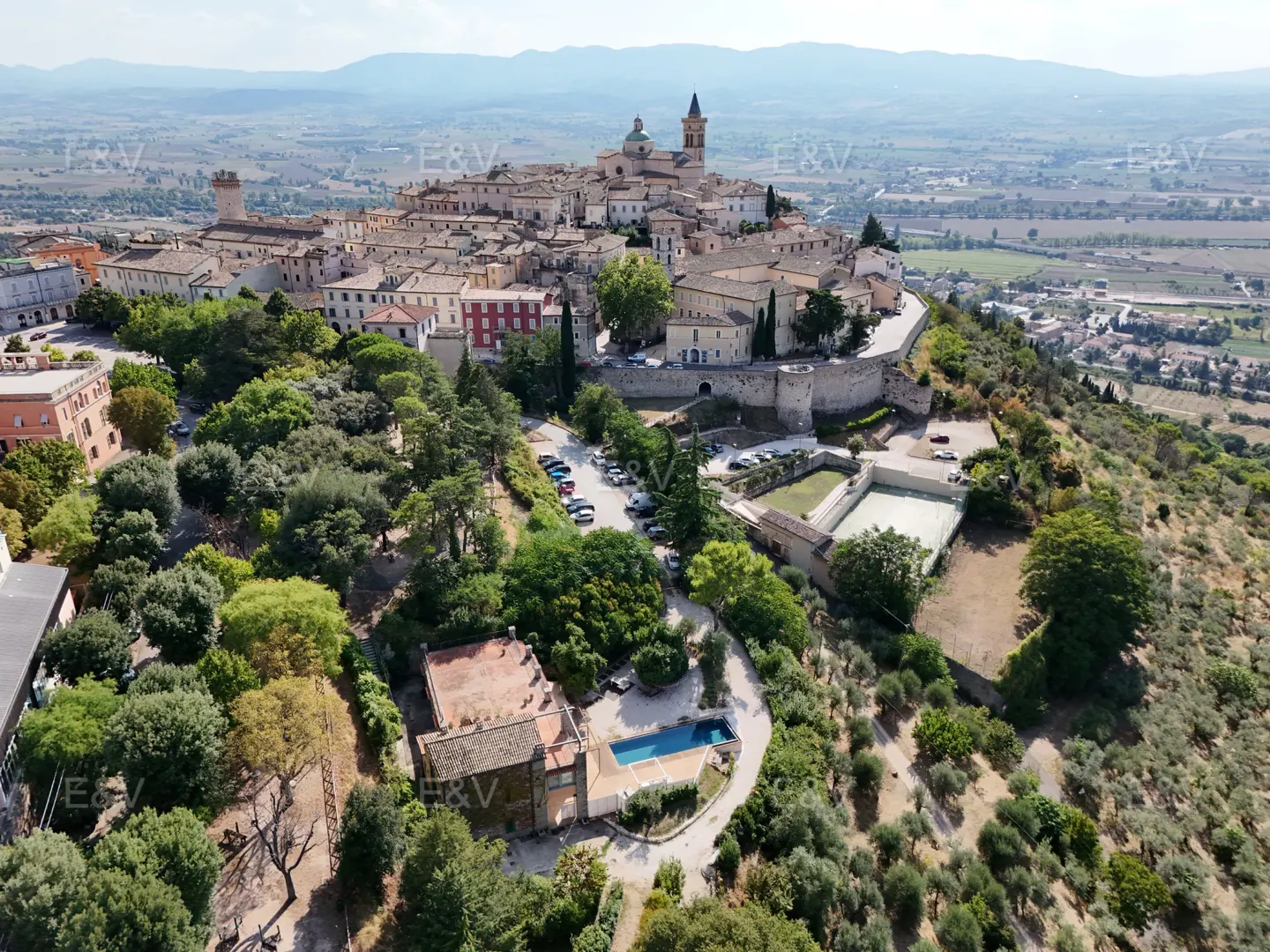Aerial view of Spello, Italy, atop a green hill. The town features stone buildings, a church steeple, and a valley in the background.