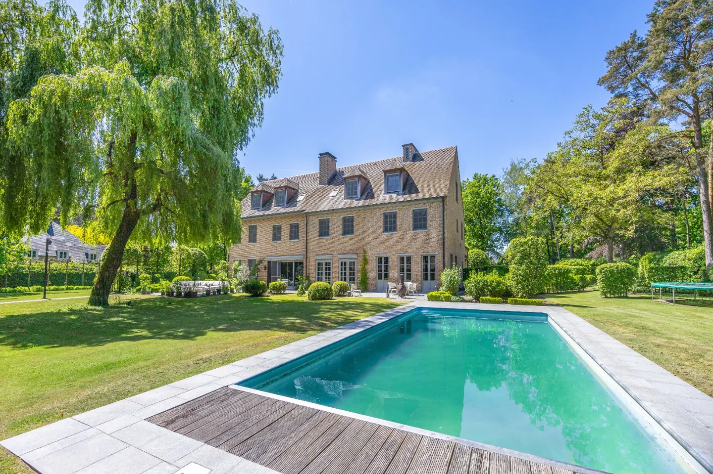 A large, tan brick house with a pool in the foreground and green trees surrounding the property.