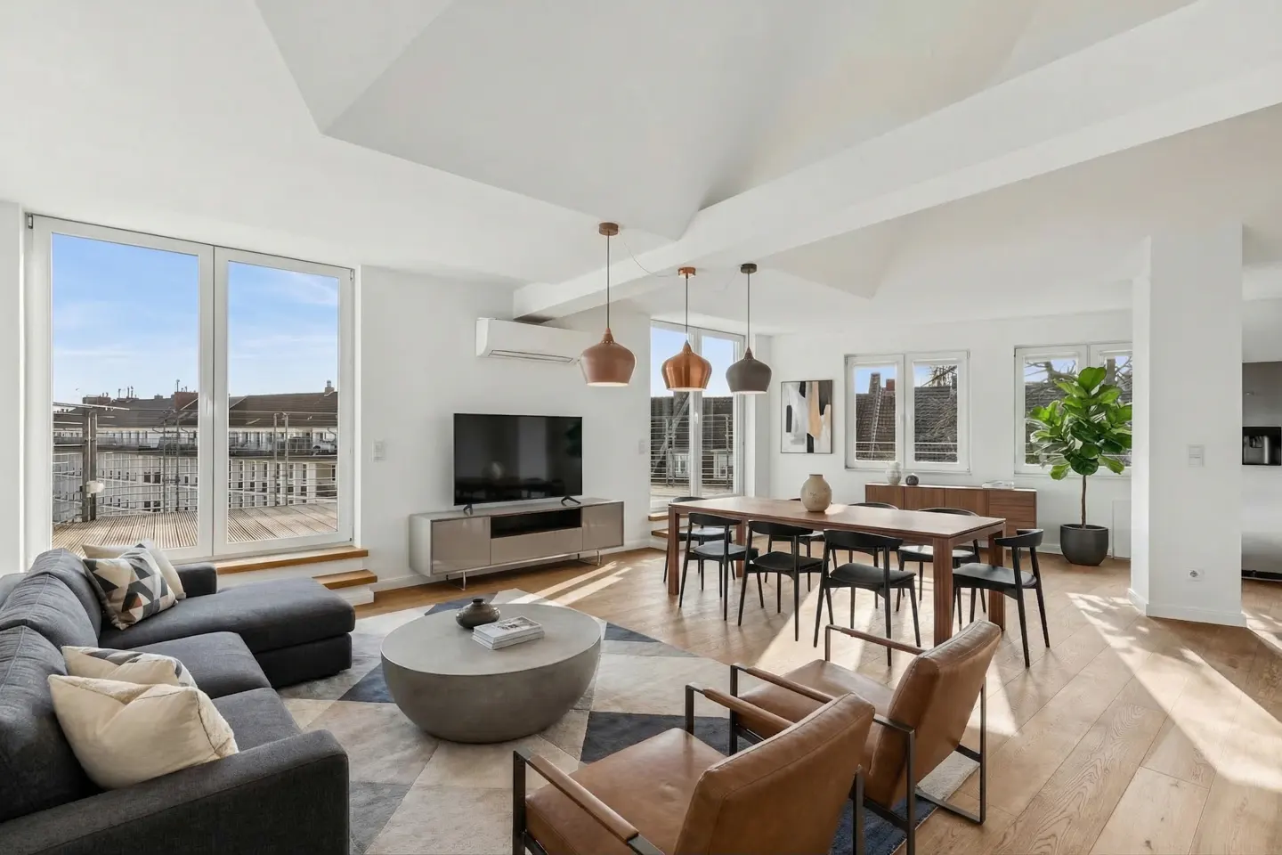 Bright, modern living room with a gray sectional sofa, round concrete coffee table, and a dining table with black chairs. Large windows offer a city view.