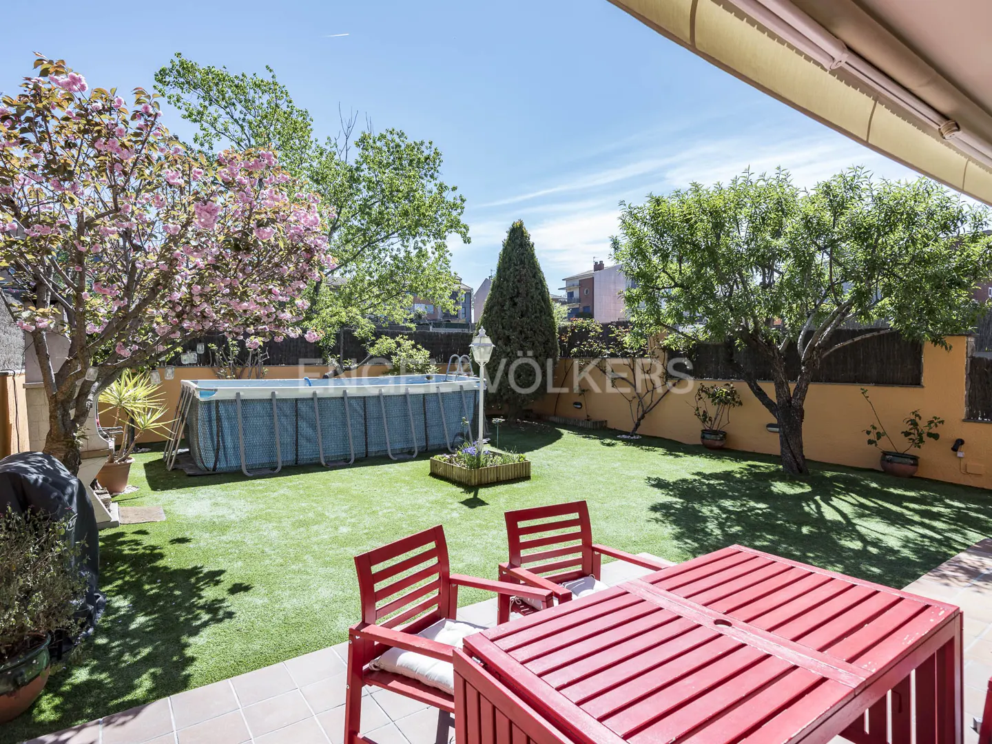 A backyard with a pool, green grass, trees, and a red table with chairs.