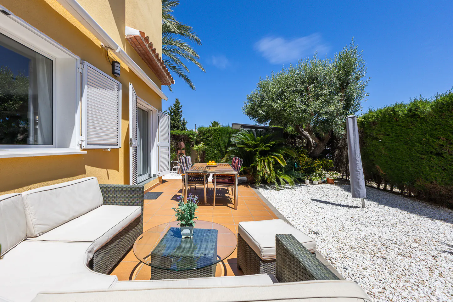 Outdoor patio with wicker furniture, glass table, and dining set on a terracotta tile floor. White gravel and green hedges in the background.
