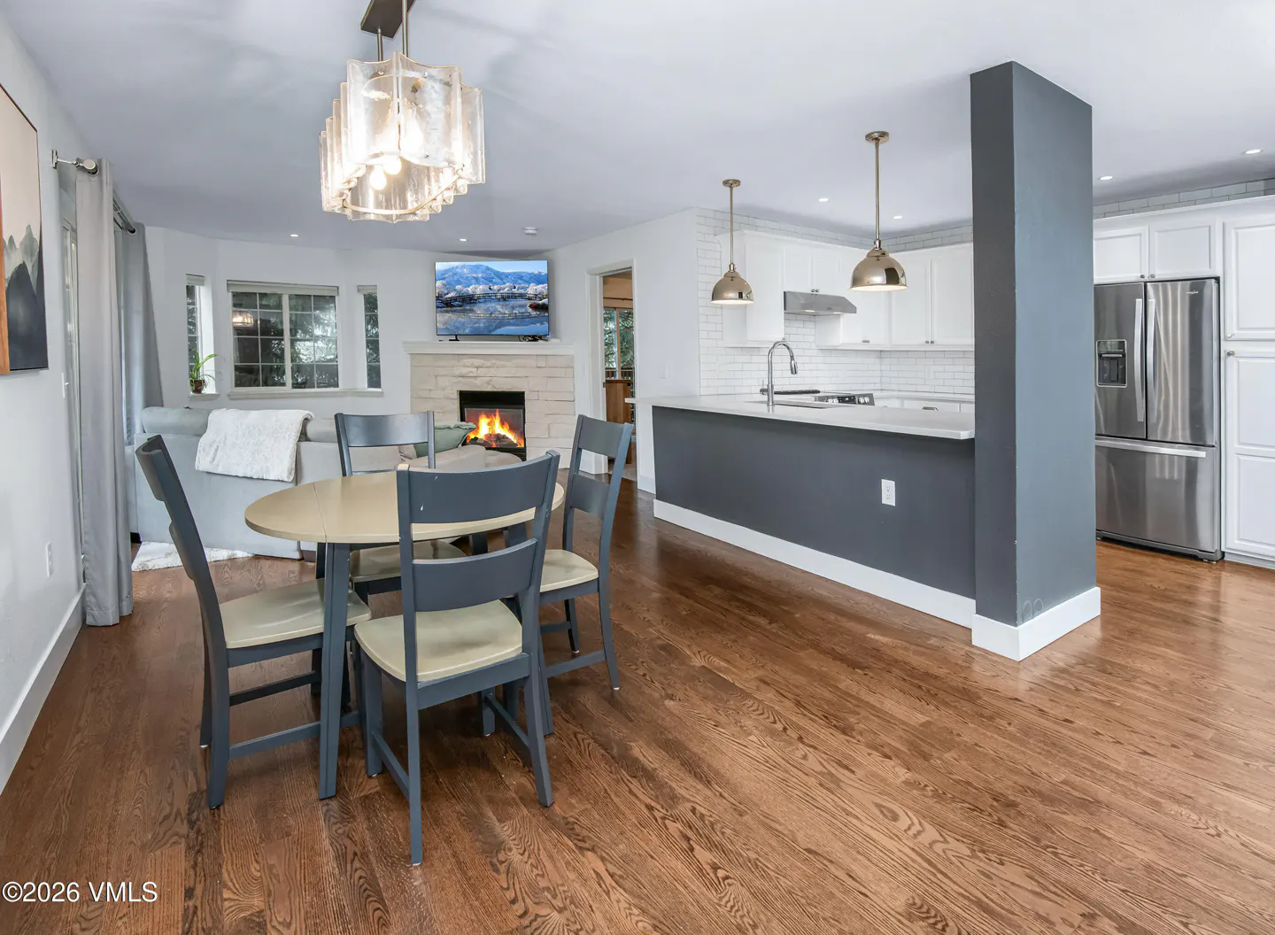Open-concept living space with wood floors, a dining table with blue chairs, and a kitchen with white cabinets and a gray island. A fireplace is visible in the background.