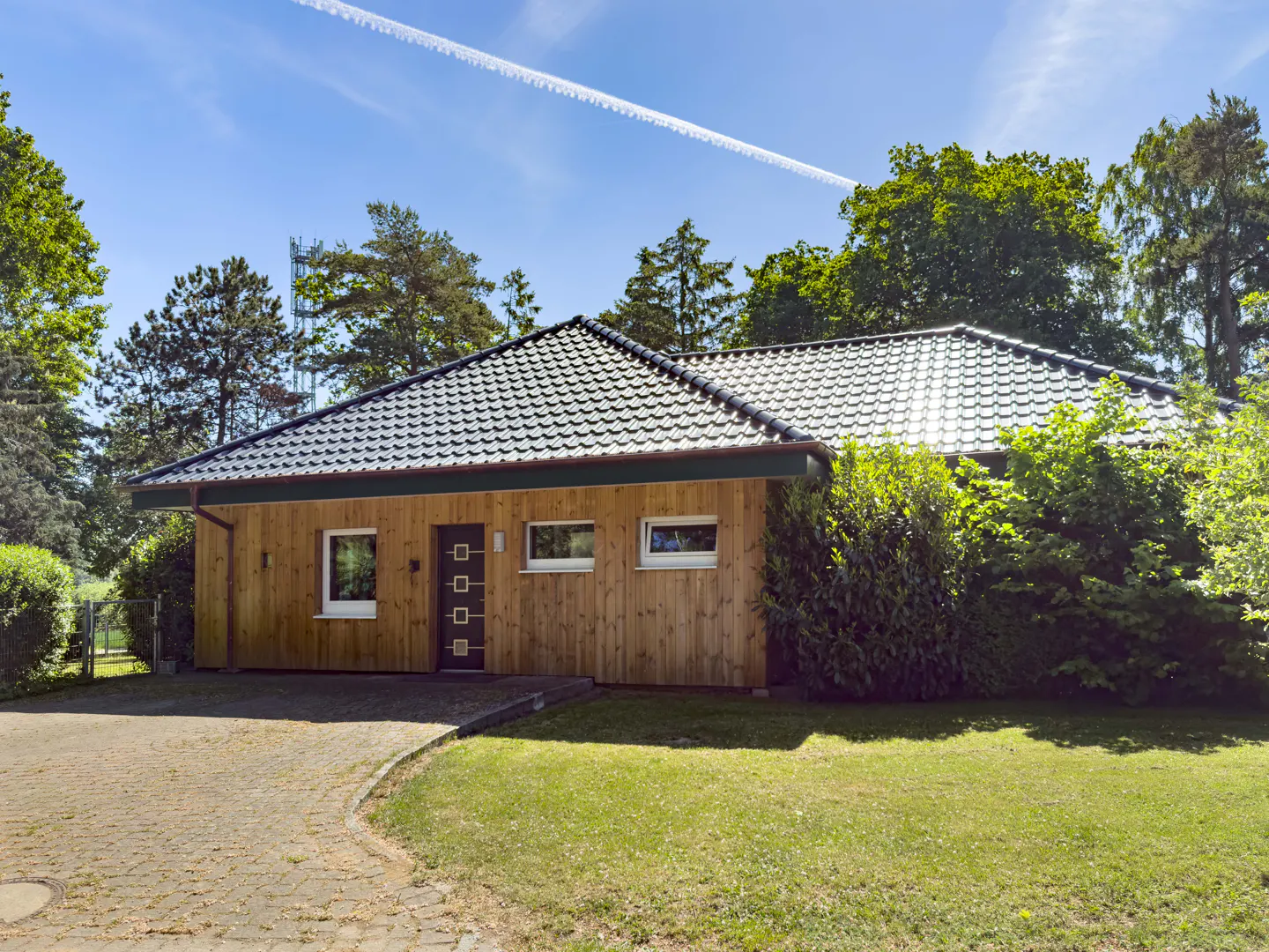 Single-story house with wood siding, dark tiled roof, and a paved driveway, surrounded by trees on a sunny day.