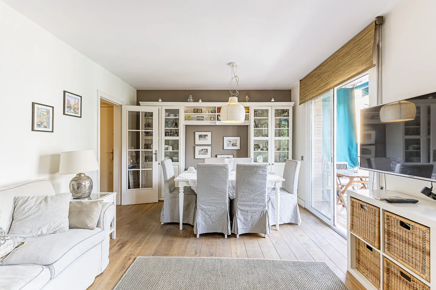 Bright living room with white sofa, dining table with grey chairs, and a cabinet filled with decorative items. Sliding glass doors lead to a balcony.