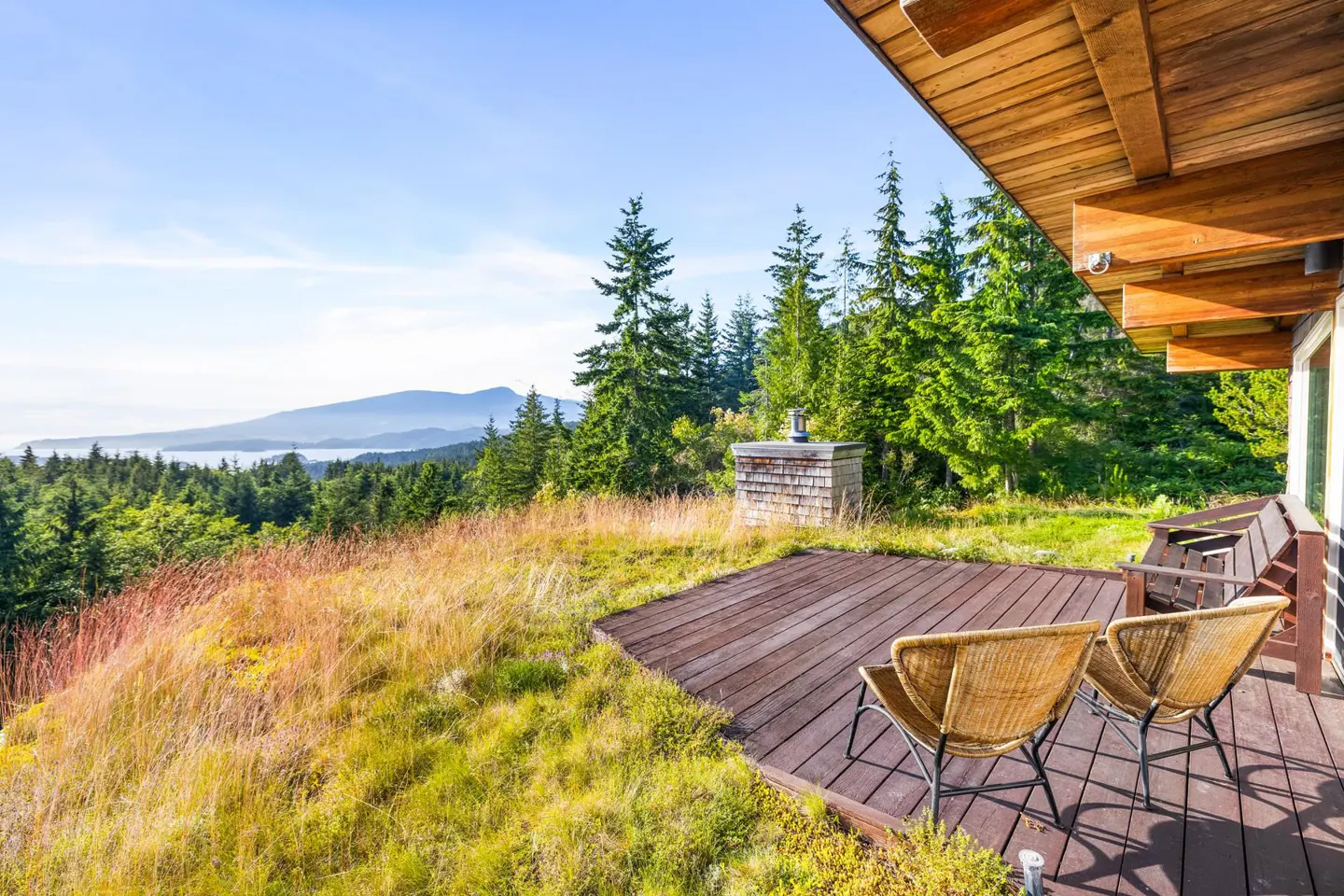 Wooden deck with two wicker chairs overlooks a grassy hillside, evergreen trees, and distant mountains under a blue sky.