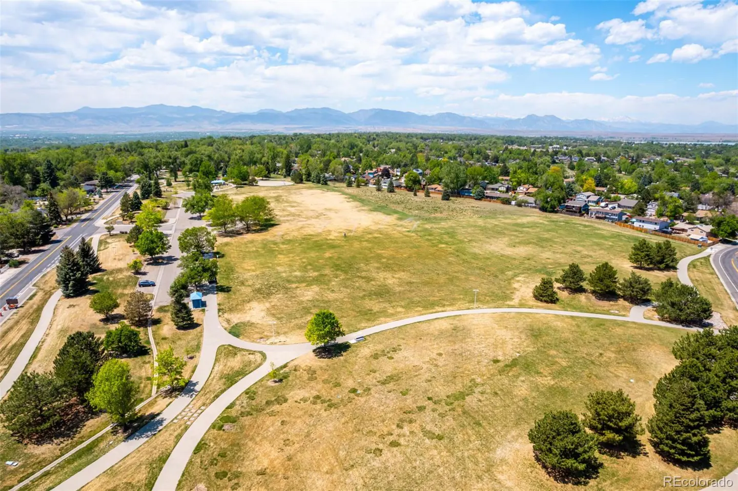Aerial view of a park with walking paths, trees, and a large grassy field. Mountains are visible in the distance under a blue sky with clouds.