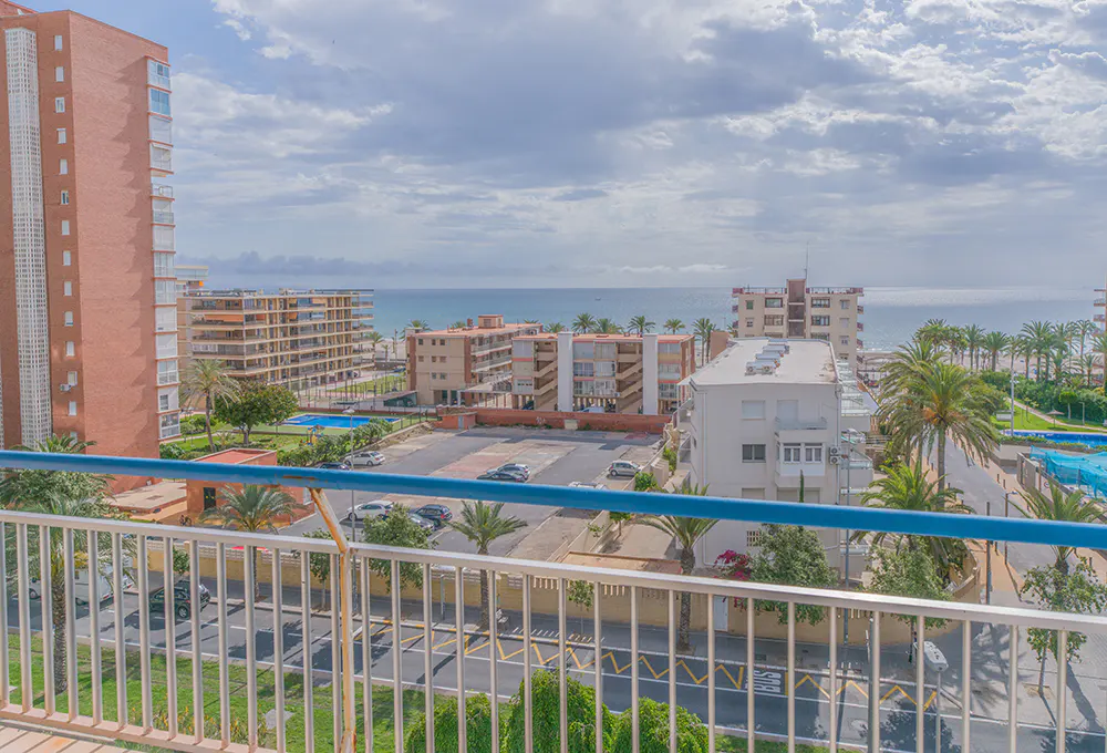 View from a balcony overlooking a coastal city with apartment buildings, palm trees, and the ocean in the background under a cloudy sky.