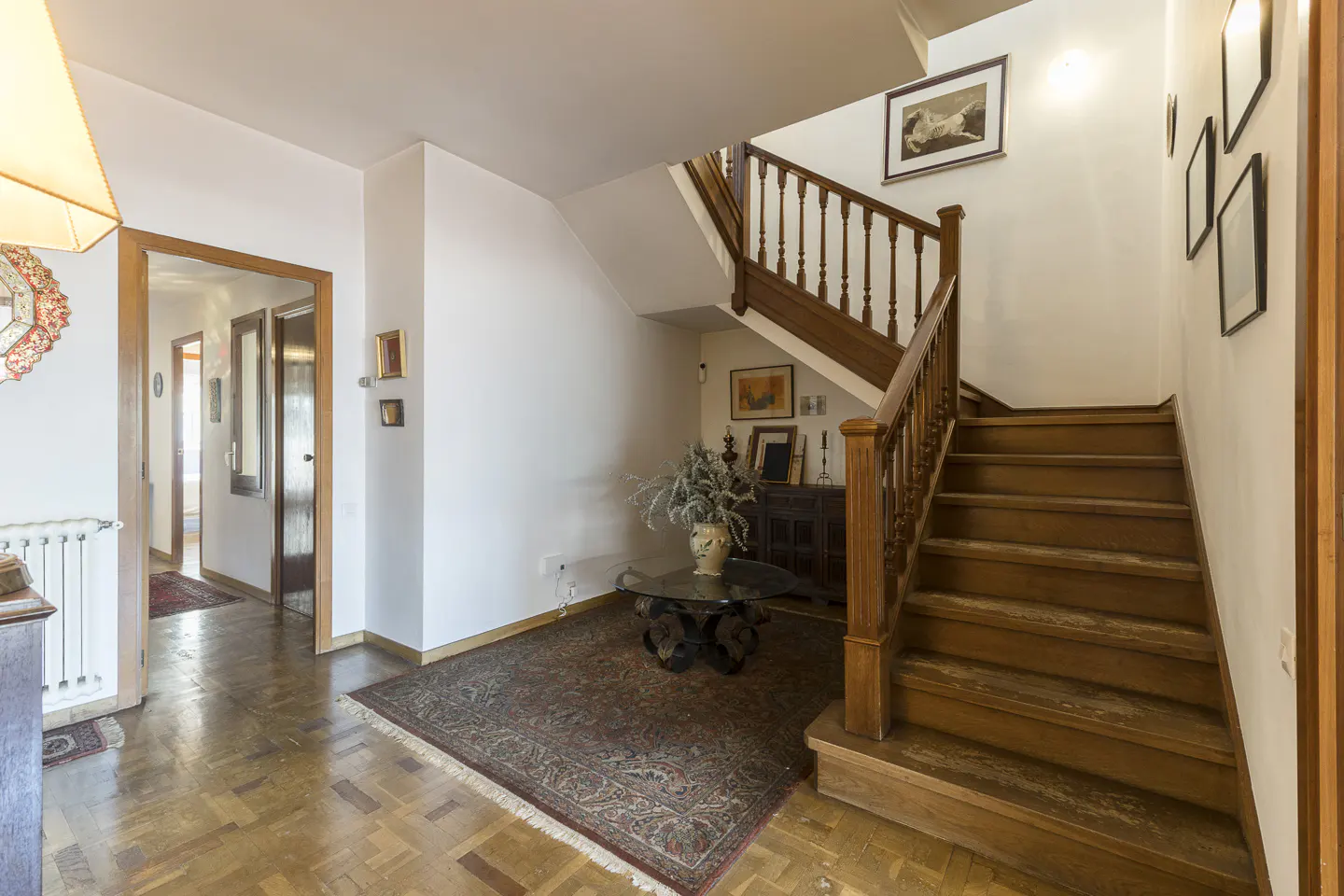 A bright foyer with wood floors, a staircase with wood railings, and a patterned rug. A glass table with a vase of flowers sits on the rug.