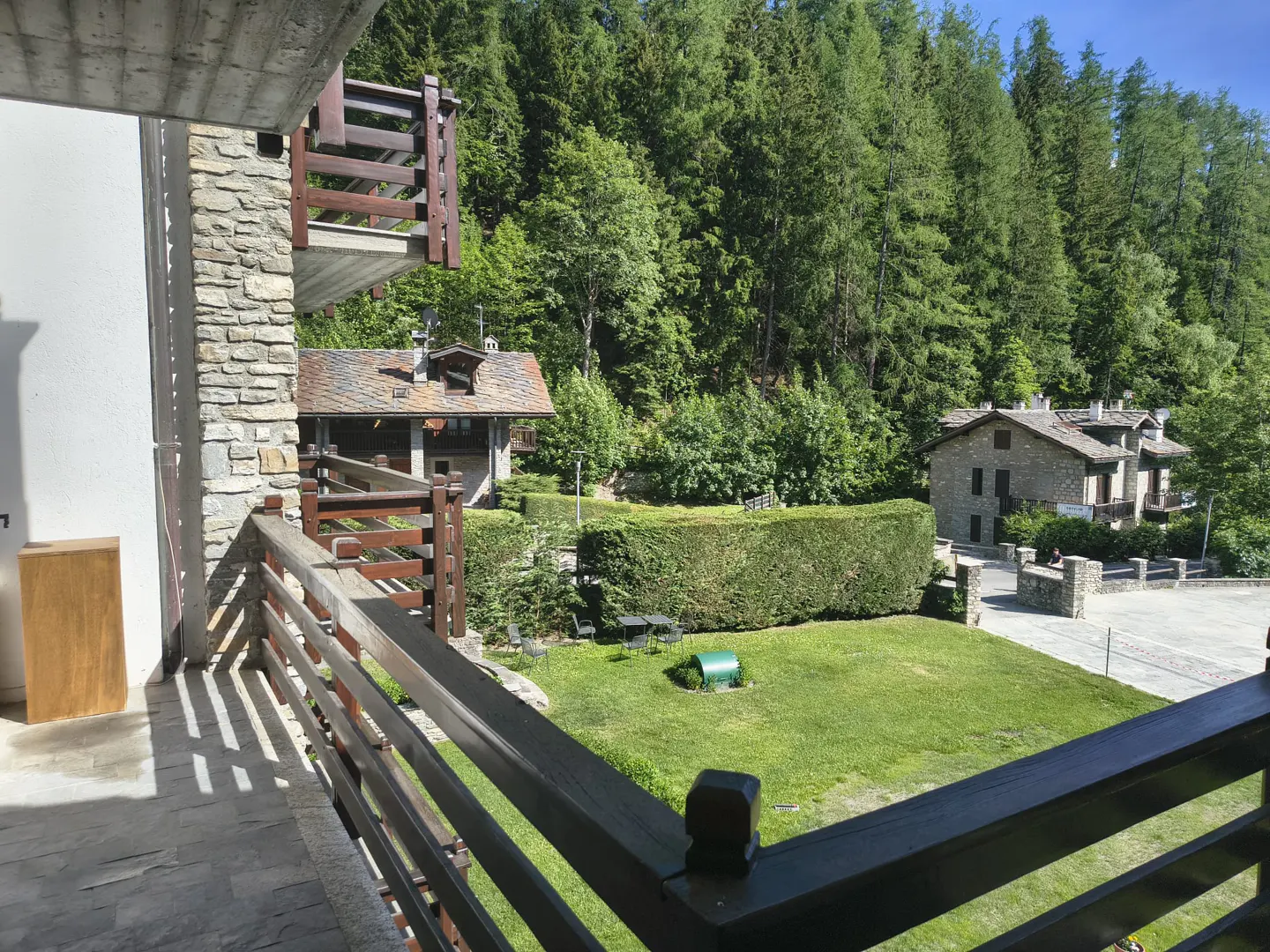 View from a stone balcony overlooking a green lawn, a manicured hedge, and rustic stone houses nestled against a forest.