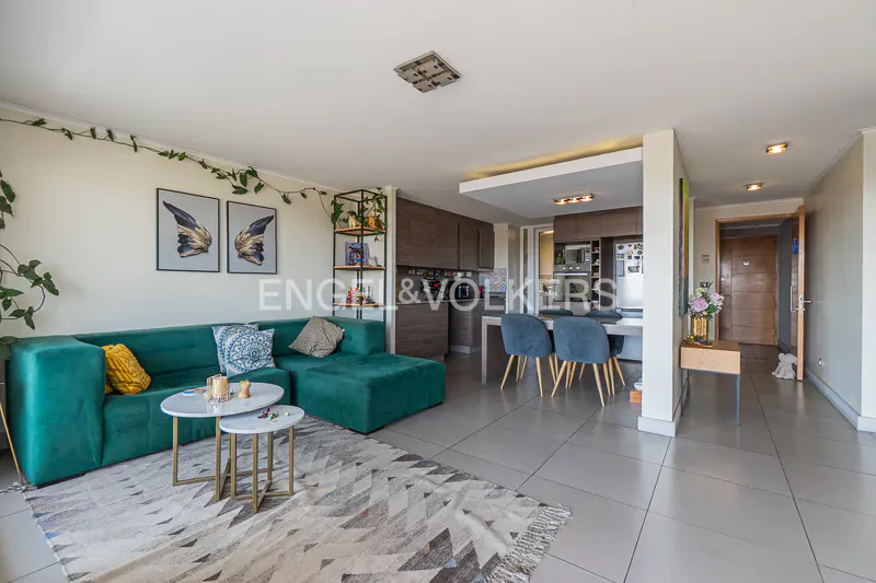 A modern apartment interior with a green sectional sofa, marble coffee tables, and an open kitchen with blue chairs.