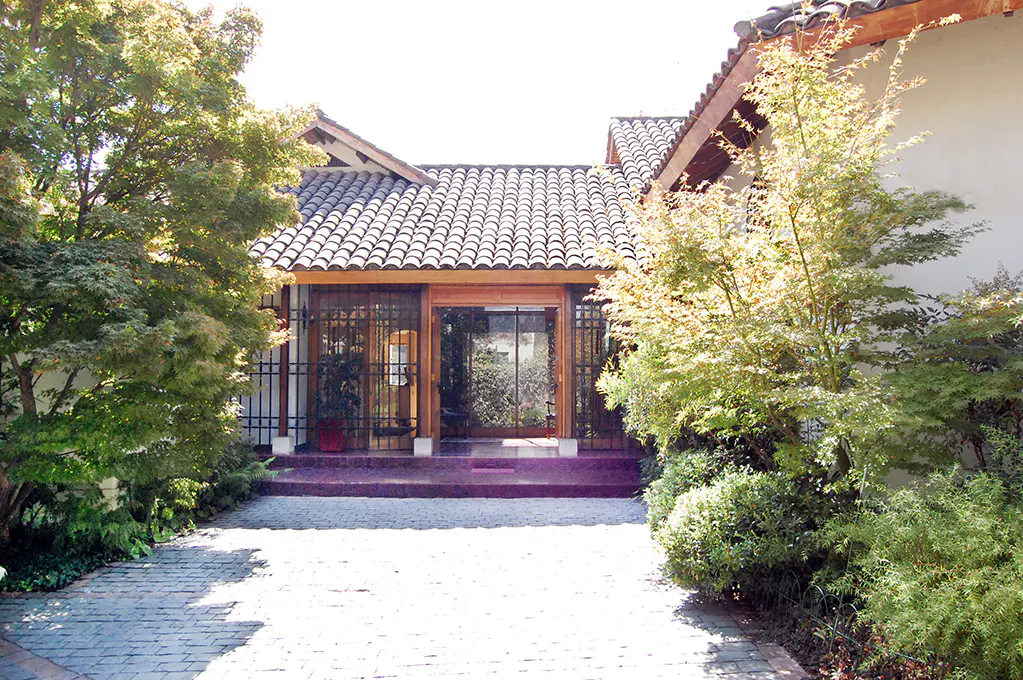 Exterior of a building with a tiled roof, wooden door frame, and lush green landscaping.