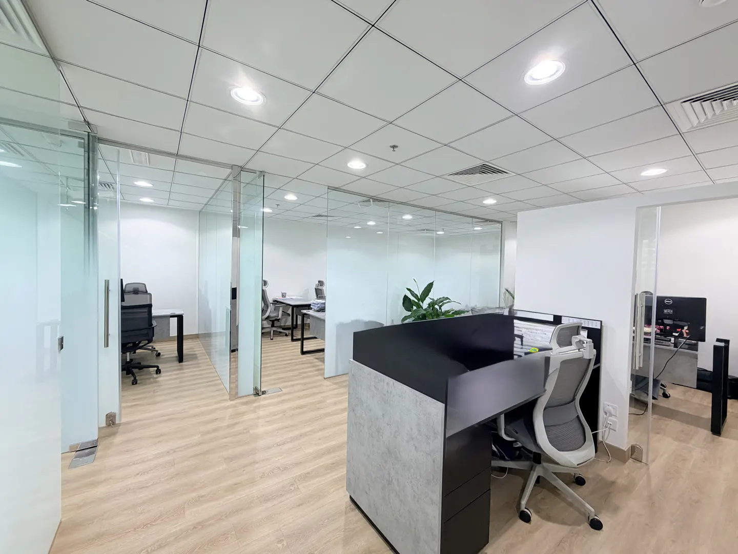 Bright office space with glass-walled rooms, desks, and chairs. Wood floors and a white ceiling with recessed lighting.