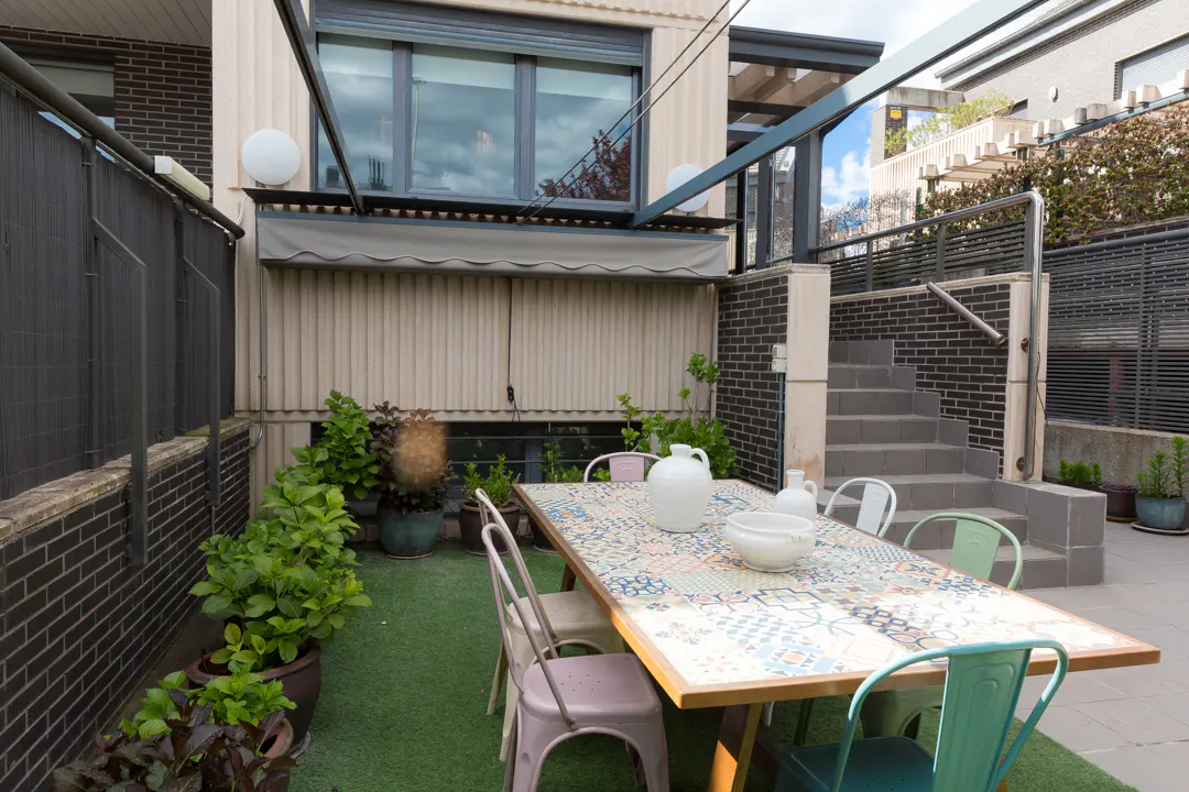 Outdoor patio with a patterned table, chairs, and potted plants on artificial grass, next to a brick building with stairs.