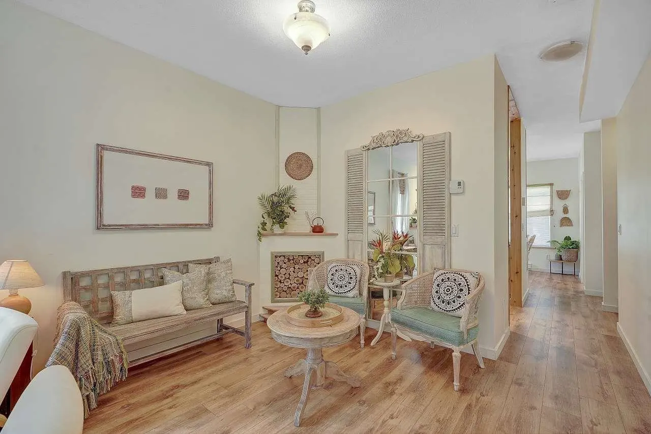 Living room with wood floors, a bench with pillows, two chairs, and a round table. A decorative fireplace with logs is in the corner.