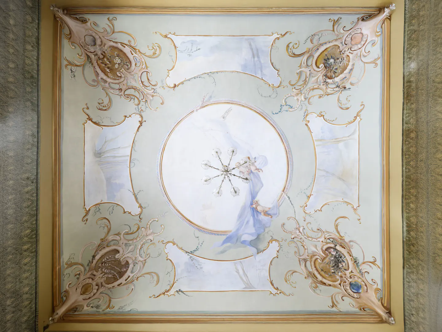 Ornate ceiling with a central chandelier and painted cherubs in blue fabric. The ceiling is framed in gold with decorative corners.