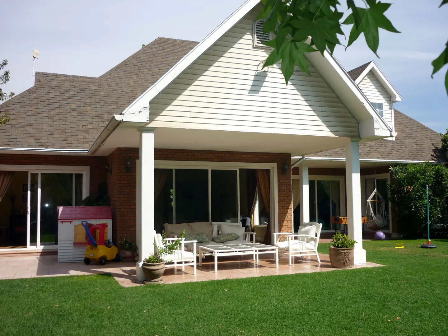 Backyard view of a brick and white siding house with a covered patio, white furniture, and green lawn.