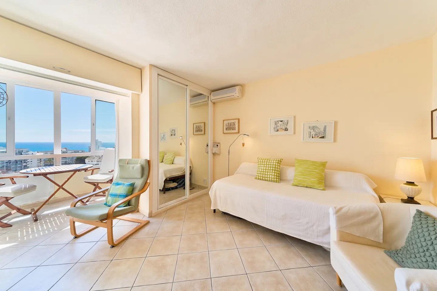 Bright studio apartment with balcony overlooking the ocean. Cream walls, tile floor, and white furniture.