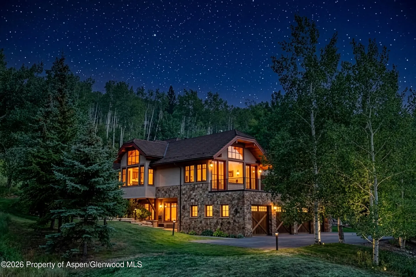 Two-story stone and stucco house at night with lit windows under a starry sky. Trees surround the house.