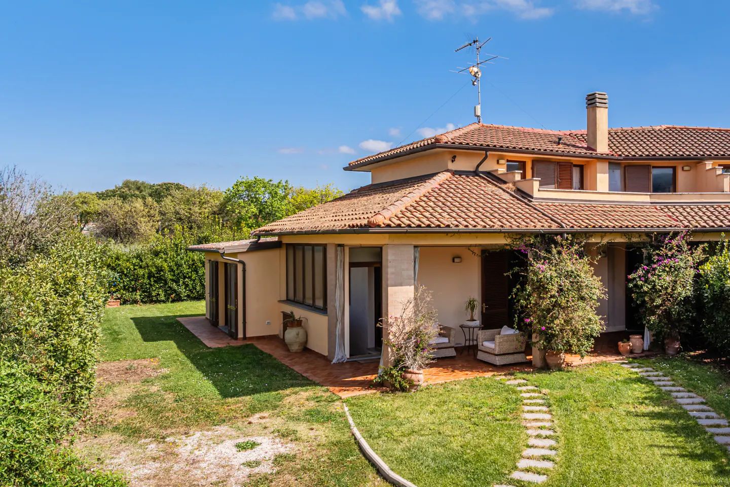 Exterior view of a tan two-story house with a red tile roof, a patio with furniture, and a stone path through the green lawn.