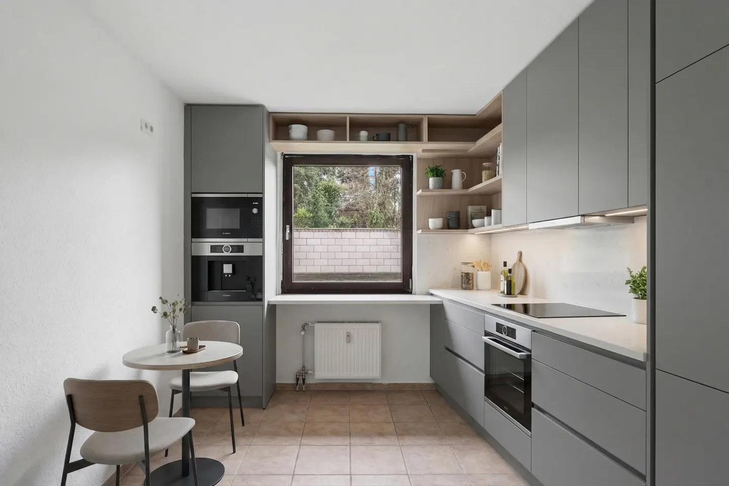Modern kitchen with gray cabinets, white countertops, and tile flooring. A small table and chairs sit near a built-in microwave and coffee machine. A window overlooks a brick wall and greenery.