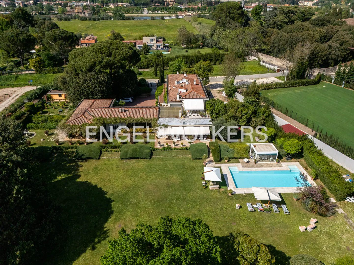 Aerial view of a large estate with a pool, lawn, and red-roofed buildings, surrounded by green trees and a field.