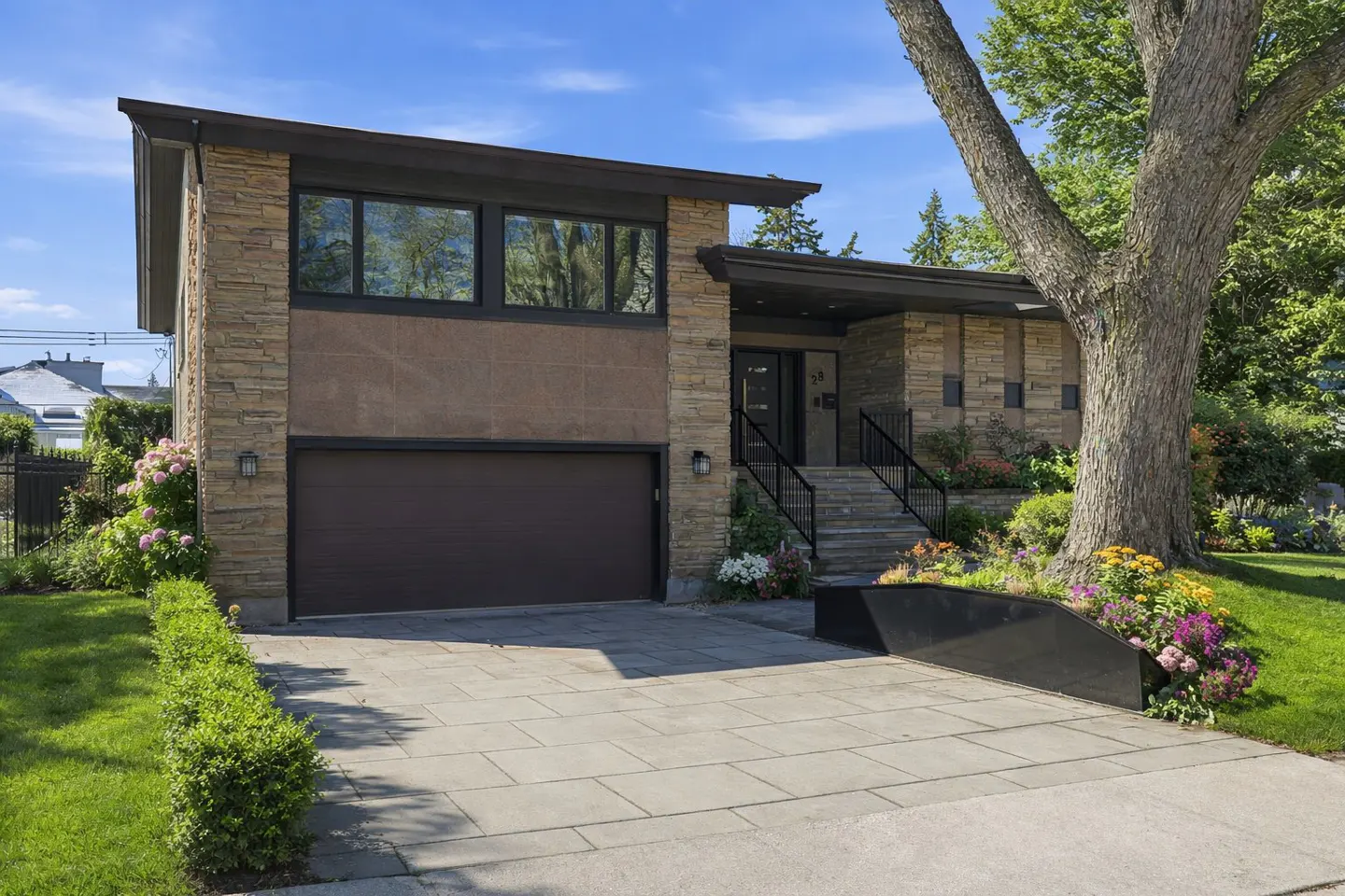 Modern stone and stucco home with a brown garage door, stone walkway, and lush landscaping under a blue sky.