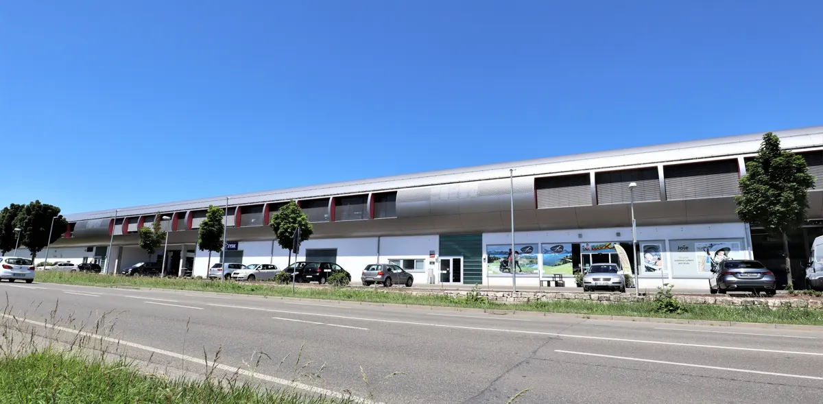 A long, one-story commercial building with a silver roof and white facade, cars parked in front, and a road in the foreground.