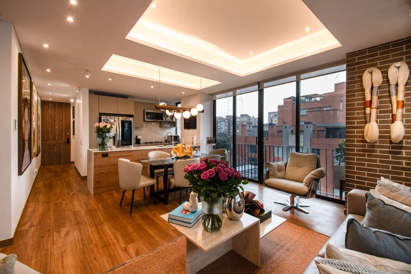 Bright, modern apartment interior with wood floors, brick accent wall, and city views. Roses on a marble coffee table.