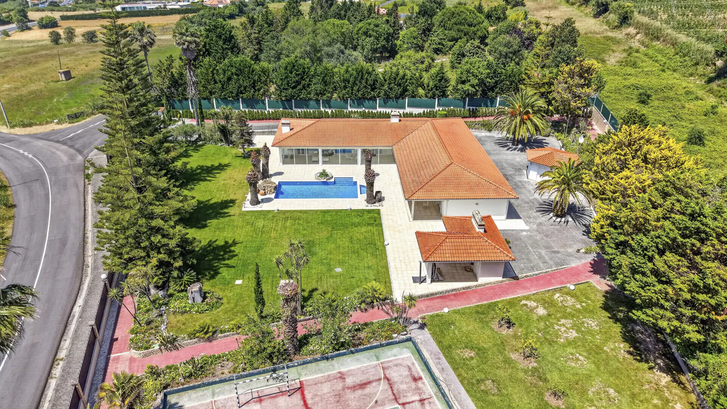 Aerial view of a villa with a terracotta roof, blue swimming pool, lush green lawn, and a private sports court.