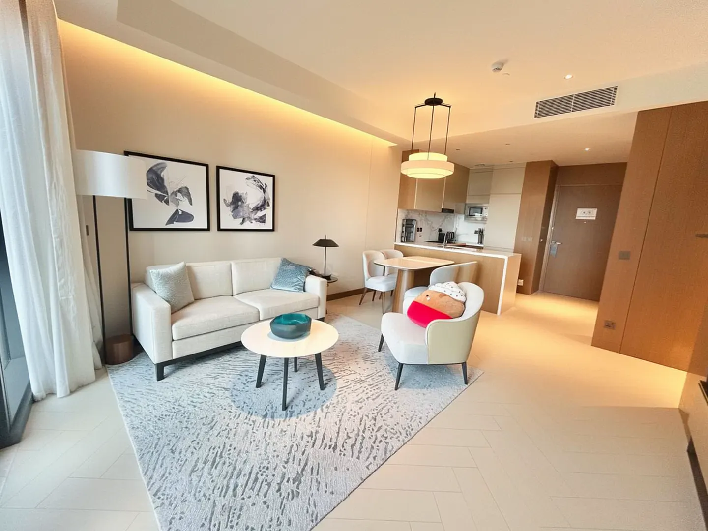 A modern apartment interior with a white sofa, round table, and armchair on a patterned rug. A kitchen and dining area are in the background.