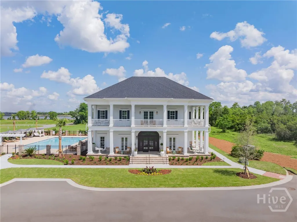 Two-story white house with black roof, columns, and balconies. Pool and trees in the background under a blue sky with clouds.