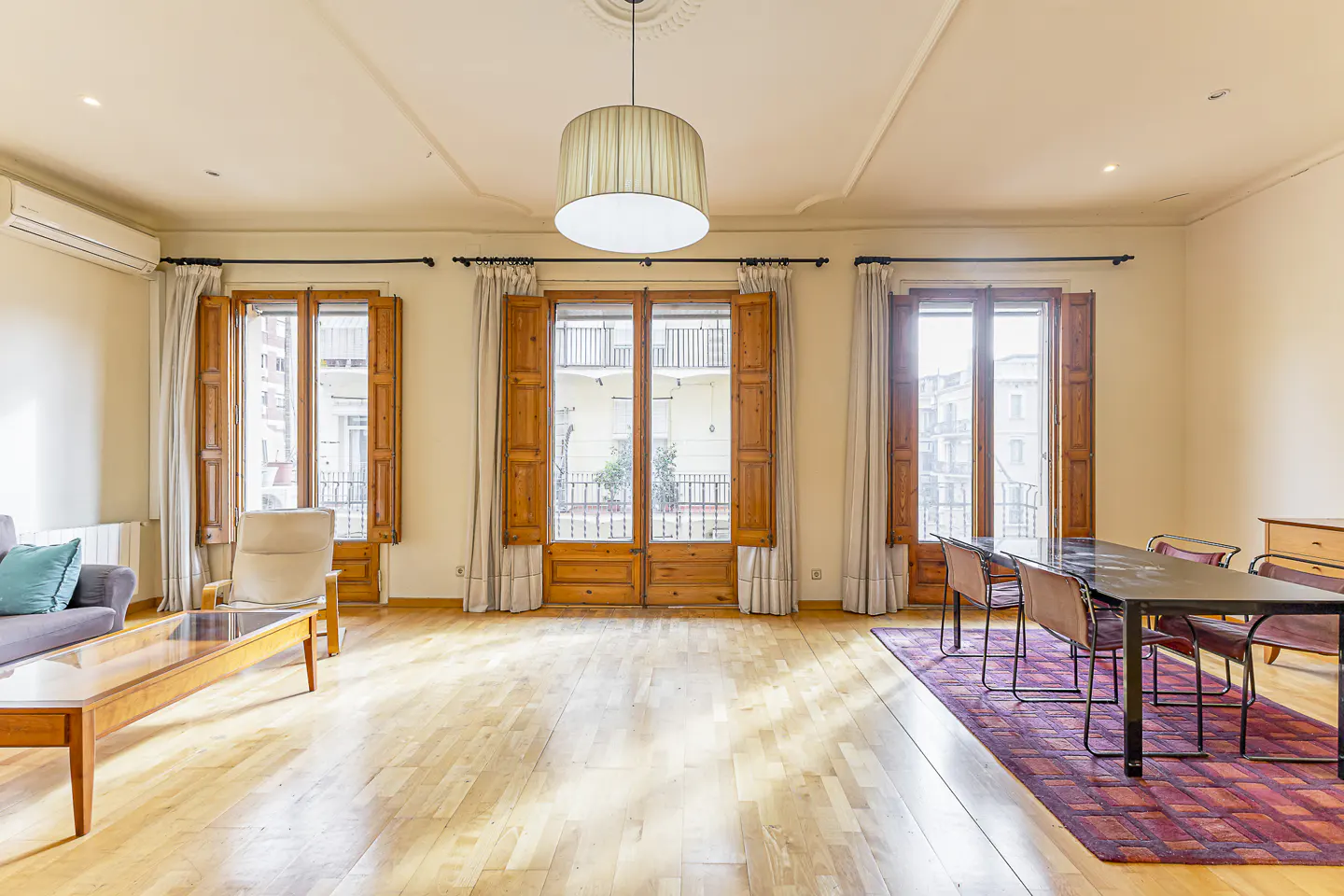 Bright living room with hardwood floors, large windows with wooden shutters, a gray sofa, and a dining table with chairs.