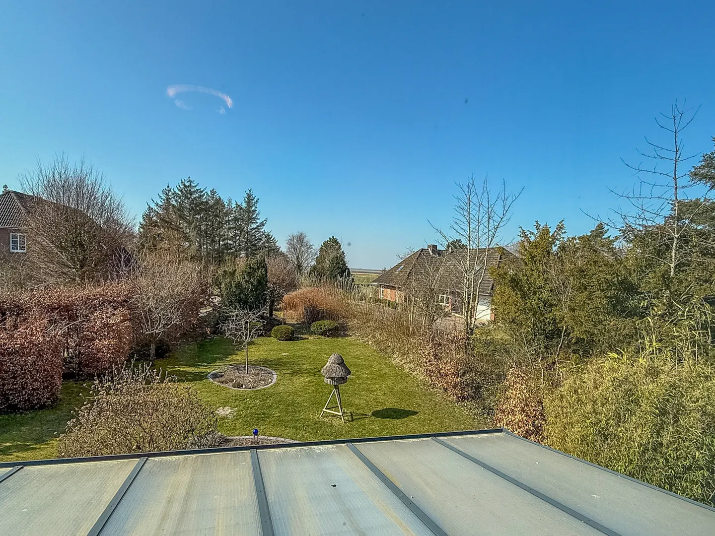 A view of a green lawn with trees, bushes, and houses under a clear blue sky. A birdhouse stands in the middle of the lawn.