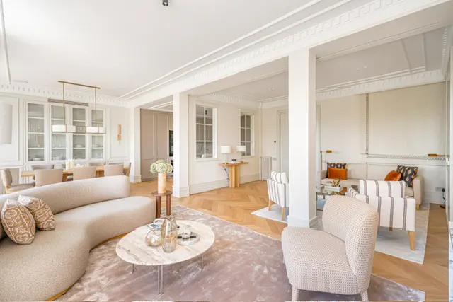 Bright, open-concept living room with beige curved sofa, marble coffee table, and herringbone wood floors. Dining area visible in background.
